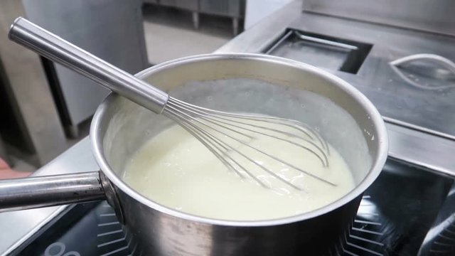 Chef stirring white bechamel sauce in a pan with metal balloon whisk. Slow motion.