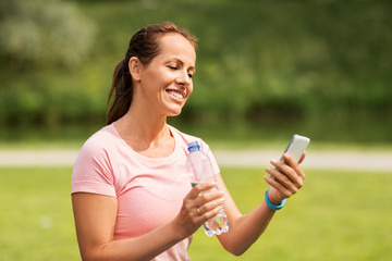 sport, technology and healthy lifestyle concept - woman with smartphone and fitness tracker drinking water after exercising in park