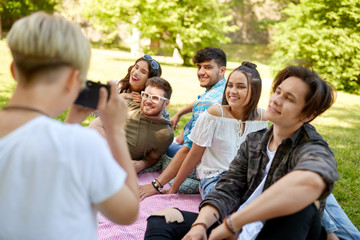 friendship, leisure and technology concept - group of happy smiling friends photographing at picnic in summer park