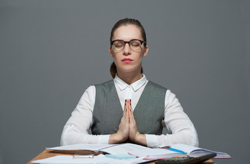 Young businesswoman dressed in a white blouse and grey jacket meditating eyes closed sitting at the desk in front of papers, taking a pause. Grey background