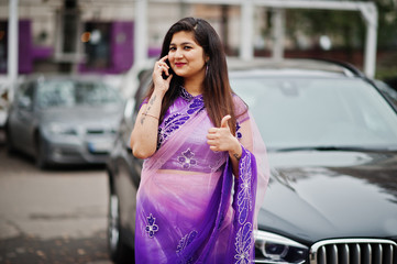 Indian hindu girl at traditional violet saree posed at street against black business suv car and speaking on mobile phone, shows thumb up.