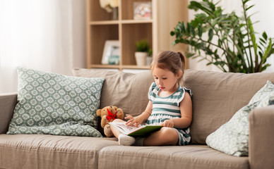 childhood and people concept - little girl sitting on sofa reading book and toy teddy bear at home