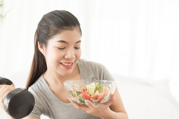 Young asian woman holding bowl of salad  with dumbell at home. Fitness and healthy lifestyle concept
