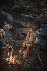 Naklejka na ściany i meble Caveman, manly boy at the fire. Scary young primitive boy outdoors near bonfire. Witch craft concept. Angry caveman, manly boy with horns near bonfire. Prehistoric tribal man outdoors on nature with