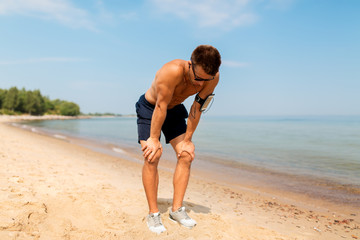Fototapeta na wymiar fitness, sport and technology concept - tired male runner with earphones and smartphone in arm band on beach
