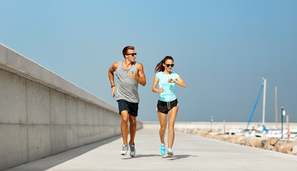 sport, technology and lifestyle concept - happy couple with fitness tracker running along pier in...