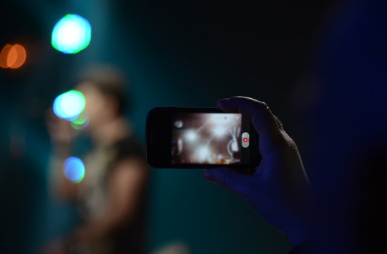 The man's hand holds the phone and takes video at the rock concert