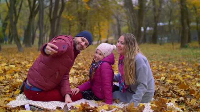 Joyful parents and smiling daughter taking selfie photo on smart phone in autumn park while sitting on picnic blanket. Happy family making self portrait with cellphone while relaxing in autumn nature.