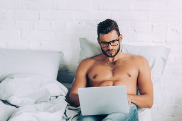 handsome shirtless male freelancer with muscular torso working on laptop in bed at home