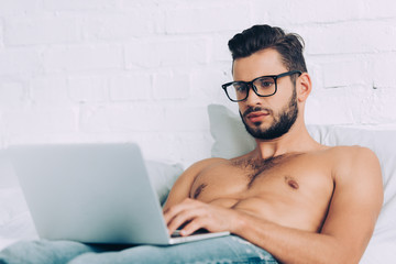 muscular shirtless male freelancer in eyeglasses working on laptop in bed at home