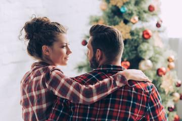 couple in checkered shirts looking at each other near christmas tree at home