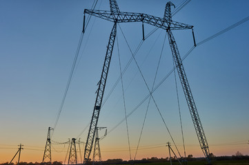 Fototapeta premium Air high-voltage transmission lines of electric energy over long distances. Overhead lines lay electricity above ground through wires attached to supports. Support, wires and blue sky at sunset