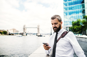 Hipster businessman with smartphone standing by the river in London, texting.