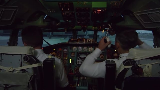 Medium shot of male pilots sitting in cockpit of airplane and preparing for flight: they are putting on headsets and pressing switch on overhead panel