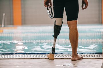 Cropped shot of swimmer with artificial leg standing in front of indoor swimming pool and holding...