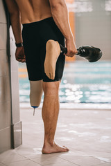 cropped shot of sportsman standing at poolside of indoor swimming pool and holding his artificial leg