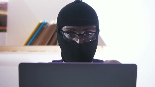 Young smart computer genius sitting at table and stealing important secret information. Portrait of masked man. Indoors. Hacker.