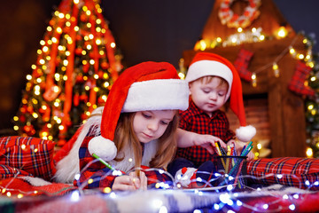 Charming girl in Santa hat lying on the floor and writes letter, draws with pencil and her little...