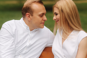 Obraz premium Handsome man look on his beautiful woman. Close up view Attractive couple sits on the bench in the park. Blonde hair man and woman spend time togerther.Background of green grass