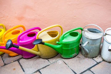 Old and used plastic and metal watering cans and pots in various colors for watering plants and flowers at home and garden stand on a stone tiles near pink rough wall. Tools for hobby and gardening