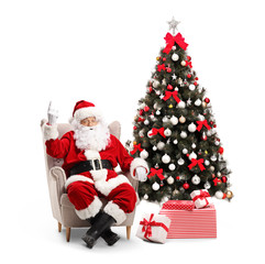 Santa Claus sitting in an armchair pointing up and a decorated Christmas tree next to him