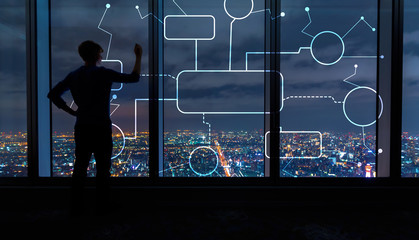 Flowchart with man writing on large windows high above a sprawling city at night