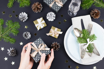 Christmas table setting with plates, presents and decorations in black and gold colors. Woman hands holding gift box