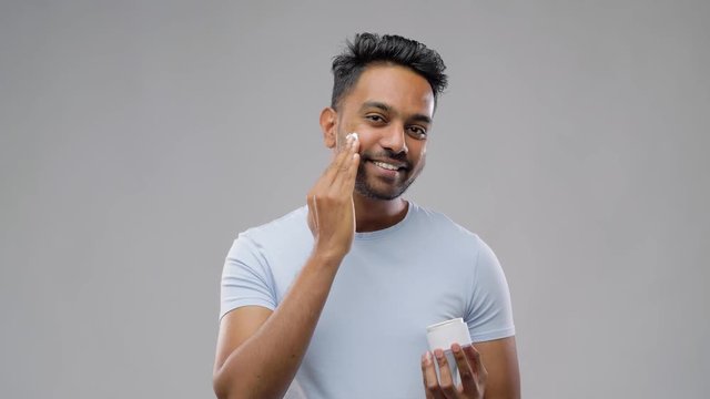 grooming, skin care and people concept - smiling young indian man applying cream to face over gray background