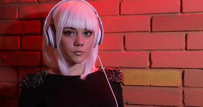 Sexy unusual girl in white wig listening to music using headphones, red light brick wall