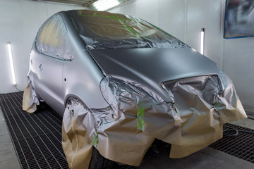 Full painting of a silver car in the body of a hatchback, some parts of which are protected by paper from splashes of paint droplets in a vehicle body repair workshop with a special box and equipment