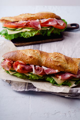 Side view of two fresh baguette sandwiches bahn-mi styled. Ham, sliced cheese, tomatoes and fresh lettuce, styled on concrete background.