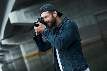 Photography hobby. Young stylish man standing on city street taking photos on camera concentrated side view