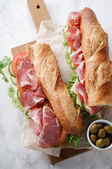 Fresh baguette sandwich bahn-mi styled. Ham, sliced cheese, tomatoes and fresh lettuce on wooden cutting board on white marble background.