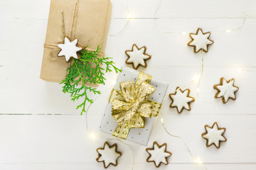 Traditional German star shaped cinnamon cookies with icing golden light garland gift box in craft and silver paper paper juniper on white plank wood background. Magic festive atmosphere. Flat lay