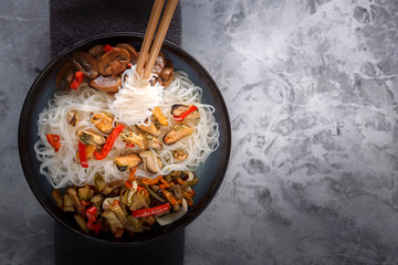 Traditional wooden chopsticks with coiled rice noodles on the background of a dish with seafood, vegetables and peppers. Copy space. Top view