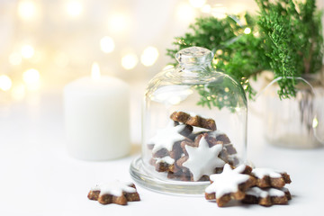 Traditional German star shaped cinnamon cookies with icing in cloche dome bell glass jar on white table. Lit candle juniper with golden garland sparkling lights. Magic festive atmosphere
