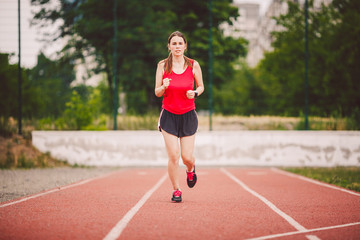 Obraz premium Beautiful young athlete Caucasian woman with big breasts in red T-shirt and short shorts jogging, running in the stadium with red rubber coating