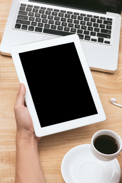 Mock up image of a hand holding black tablet pc with white blank screen and coffee cup on wooden table background