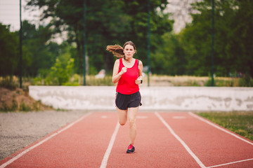 Obraz premium Beautiful young athlete Caucasian woman with big breasts in red T-shirt and short shorts jogging, running in the stadium with red rubber coating