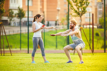 Team work in sports. A female trainer is training a young handsome man with a beard in a park on green grass, a lawn at sunset. A girl holds a phone in her hand and uses a stopwatch