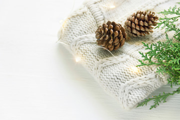 Fototapeta na wymiar Folded knitted off-white wool sweater small pine cones green juniper twig golden lights garland on plank wood table by window. Cozy winter evening. New Year Christmas magic atmosphere. Scandi style