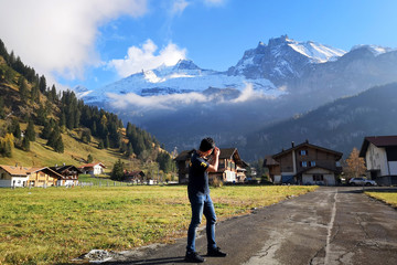 Fototapeta na wymiar Traveler is standing and taking photo of landscape view of Alps mountain and village in Kandersteg, Switzerland. Seen from his back in nice blue sky day. Travel in Europe concept.