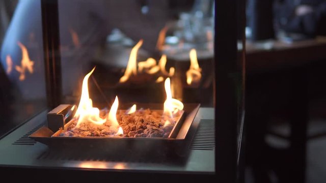 Stone Gas Fire Pit With Flames In Modern Fireplace In The Restaurant With Blurred People Meeting At Dining Table In The Background