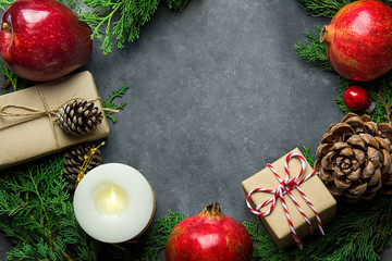 Festive colorful botanical Christmas composition. Fresh green juniper red apples pomegranates pine cones bauble candle gift boxes in craft paper arranged in wreath on dark stone background