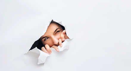 A girl with a cunning expression. Portrait of a woman looking through the hole in white paper. Distrustful look. Women's curiosity and gossip. A jealous wife.