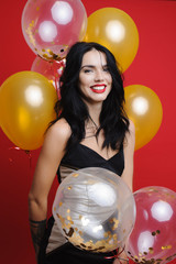 Naklejka premium Charming young female in evening dress cheerfully smiling and looking at camera while standing on bright red background and holding bunch of balloons.Elegant woman with balloons