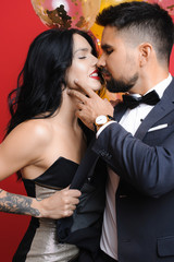 Side view of sensual woman keeping eyes closed and grasping elegant jacket of handsome man before kissing him near balloons on red background. Woman grasping jacket of man before kiss