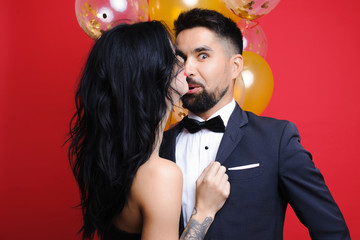 Elegant lady kissing amazed guy in nose while standing near bunch of balloons on red background. Woman kissing surprised man near balloons