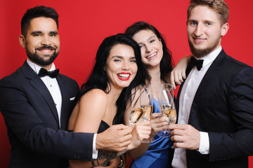 Group of attractive young people in elegant outfits holding glasses of champagne and laughing while standing on vibrant red background. Young people with champagne laughing