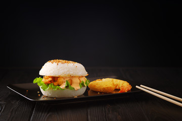 Fusion food, japanese style rice burger with tempura prawns on a black plate on black wooden background.